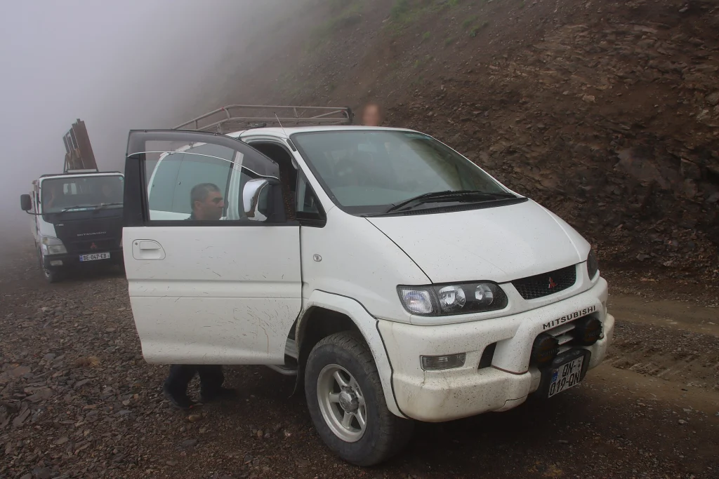 A mud-splattered 4x4 minivan navigates the gravel track over Abano Pass in misty weather.