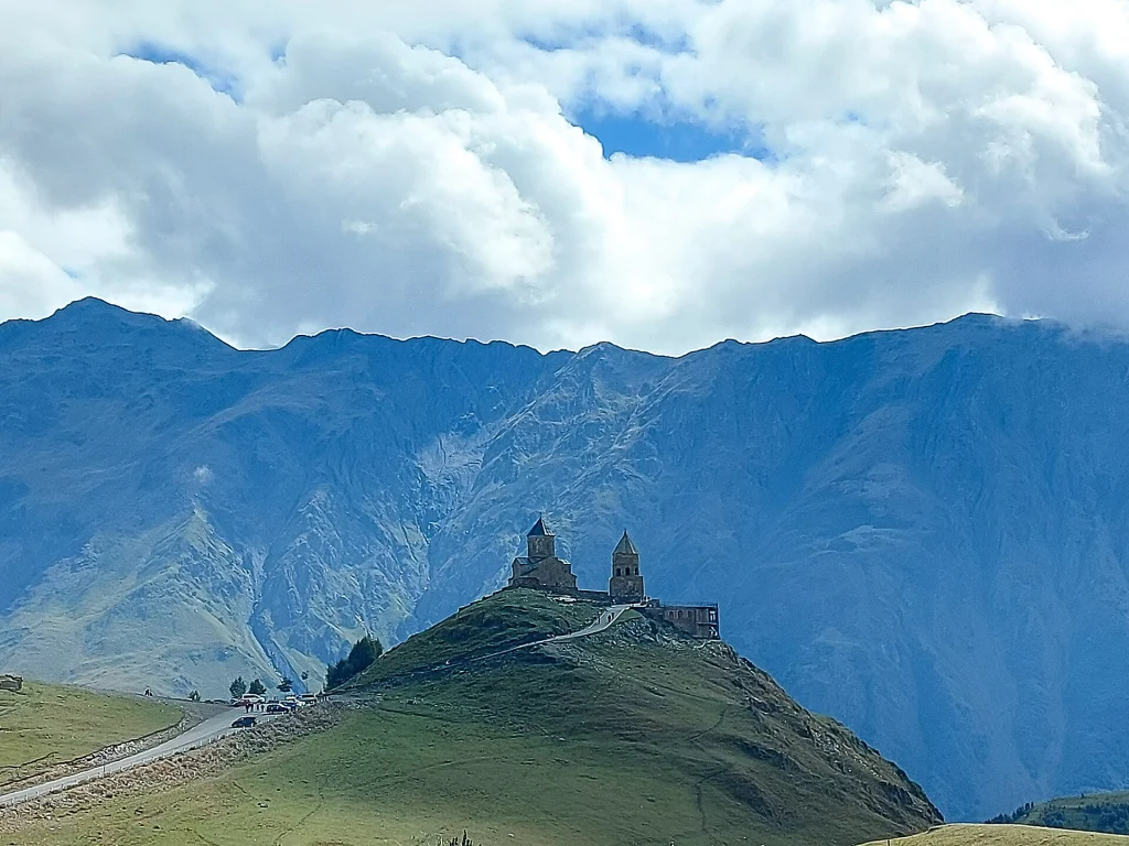 Gergeti Trinity Church perched on a green hilltop beneath dramatic Caucasus mountains and clouds.