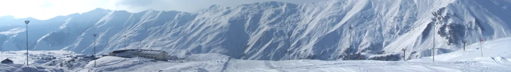 Wide panoramic banner of Gudauri's snowy peaks with ski lift infrastructure.