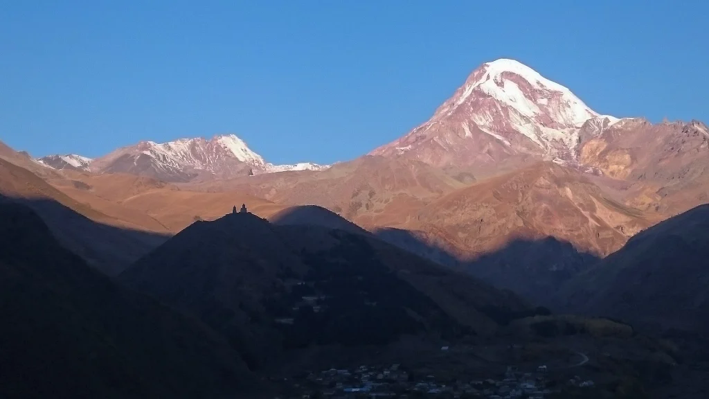 Kazbek mountain lit by morning sun with Gergeti Church shadowed in the valley below.