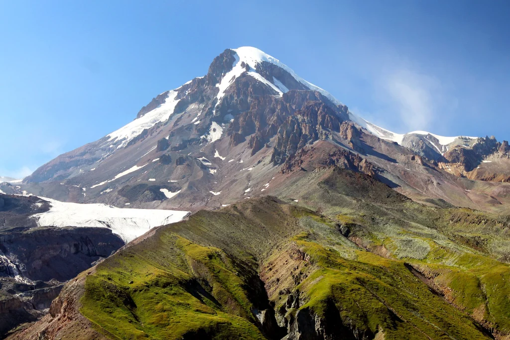 Mount Kazbek's snowy summit with the glacier and green ridges in the foreground.