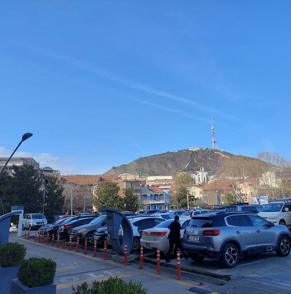 Cars fill a paid parking lot near Tbilisi's Public Service Hall, with Mtatsminda hill behind.