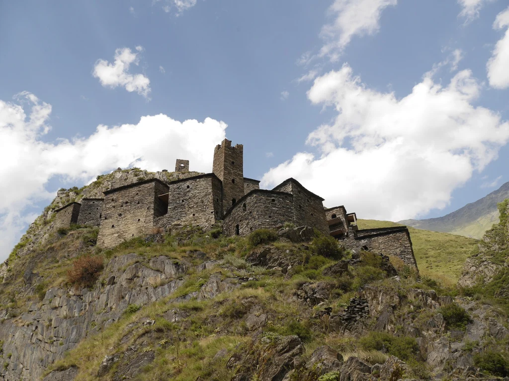 Stone towers and dwellings of Mutso fortress cling to the cliffside beneath a bright sky.