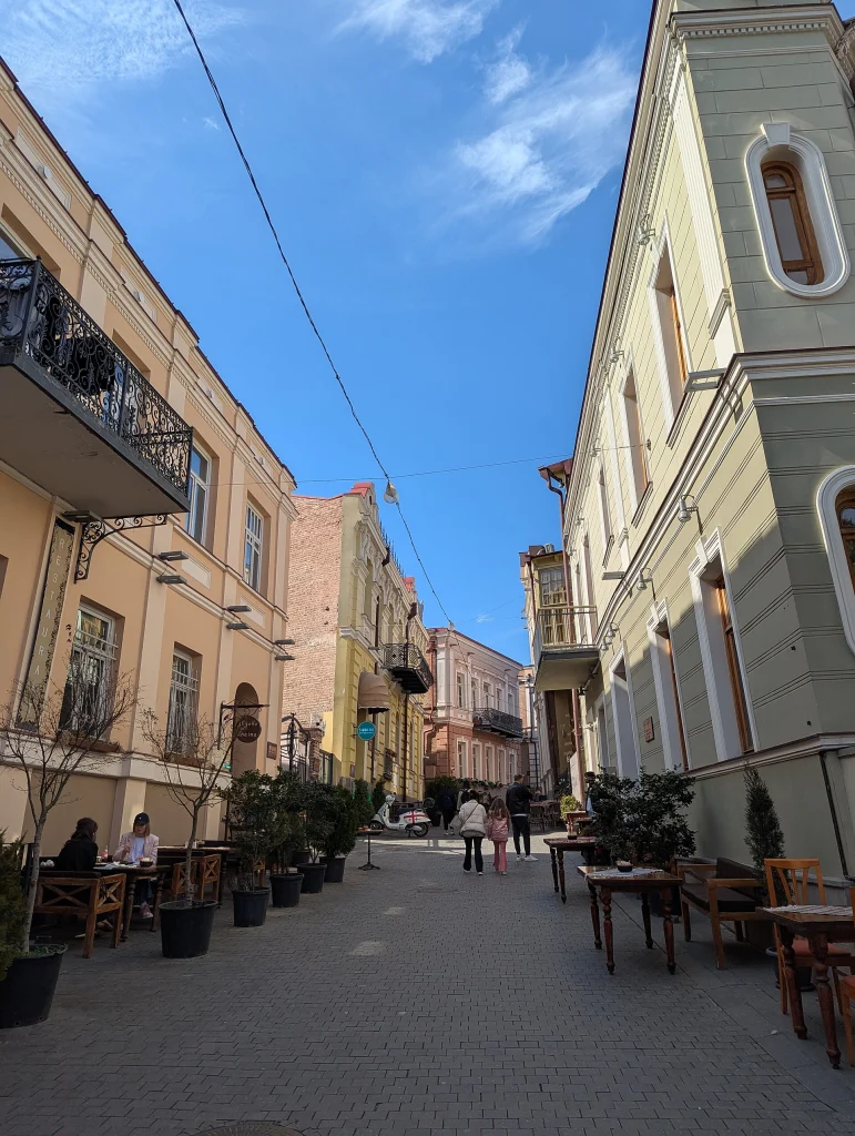 A quiet pedestrian lane in old Tbilisi with outdoor café seating, typical of mild March days.