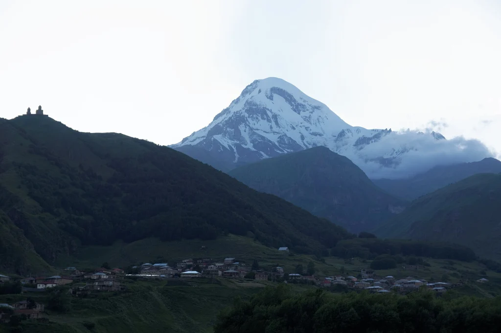 Mount Kazbek's snowy peak towers over the Gergeti valley at dusk.