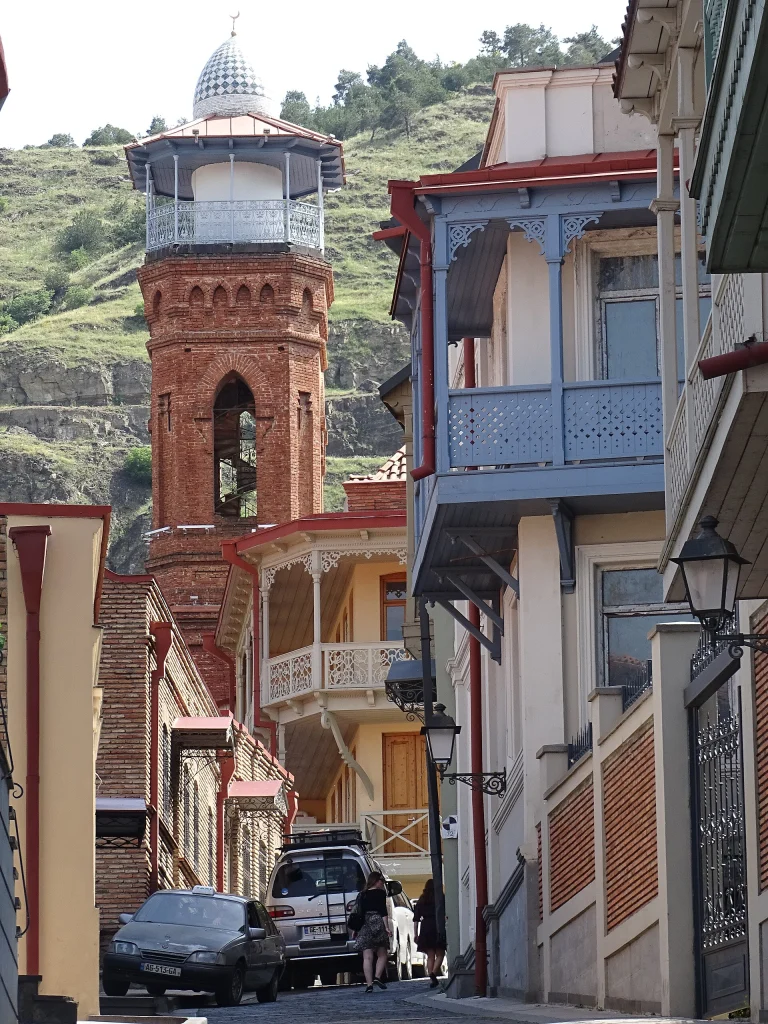 A cobbled street in Tbilisi's Old Town lined with carved balconies and a historic brick tower.