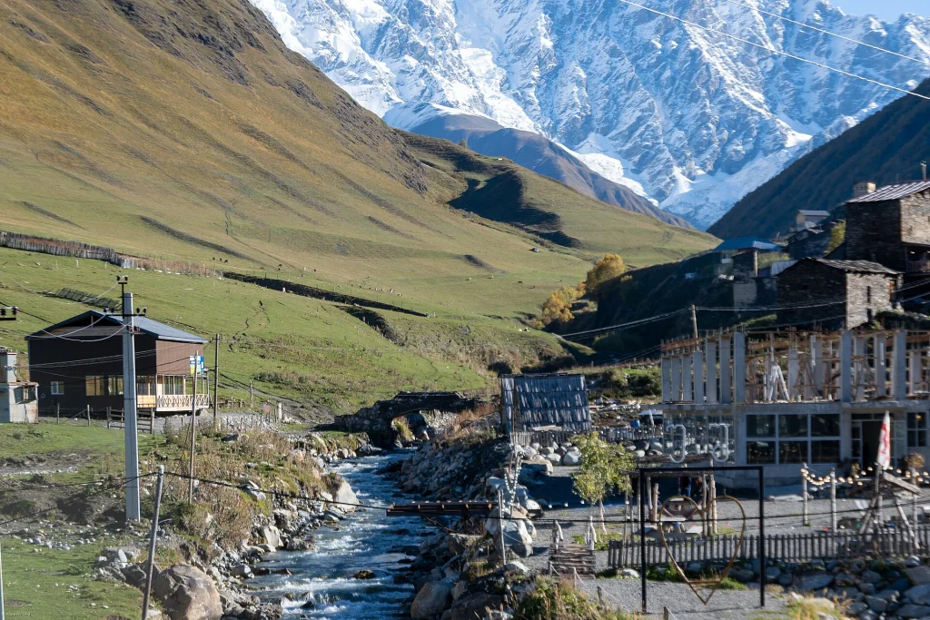 The village of Ushguli sits in a green valley with a mountain river and snowy Caucasus peaks beyond.
