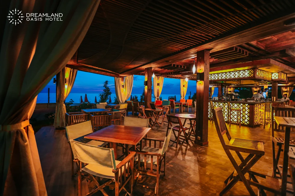 Beach Bar — Covered terrace restaurant with wooden tables and a sea view at dusk.