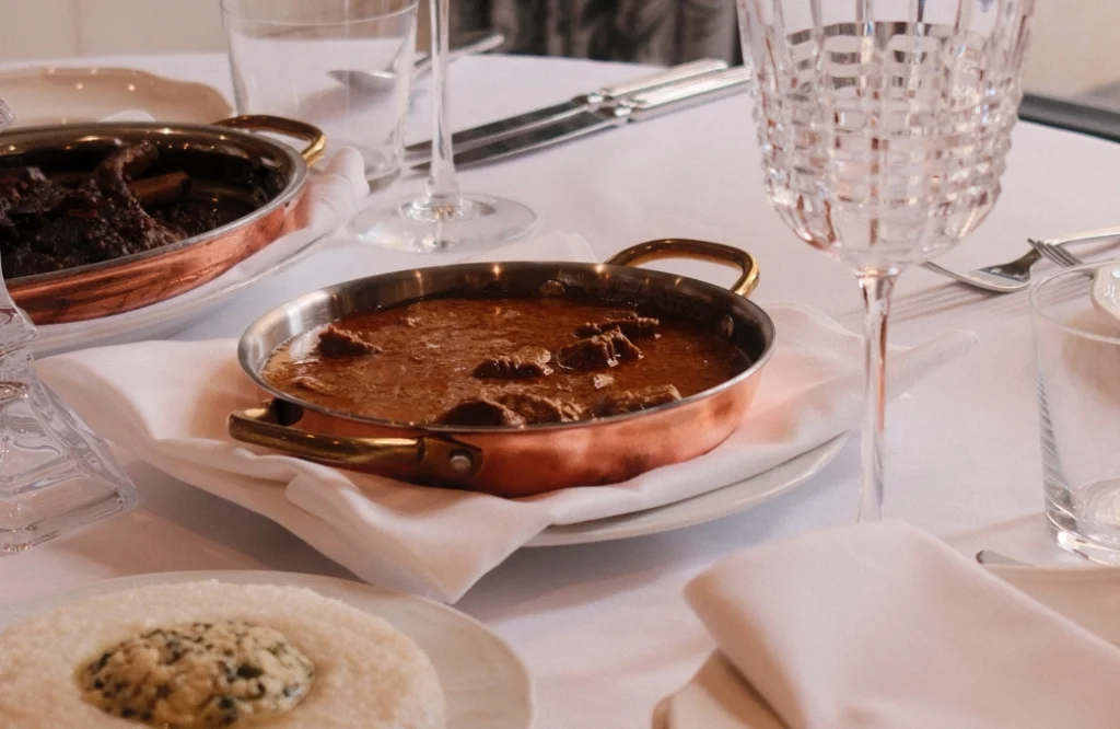Daira Restuarant — Copper pan of braised meat served tableside alongside wine glasses and formal place settings.