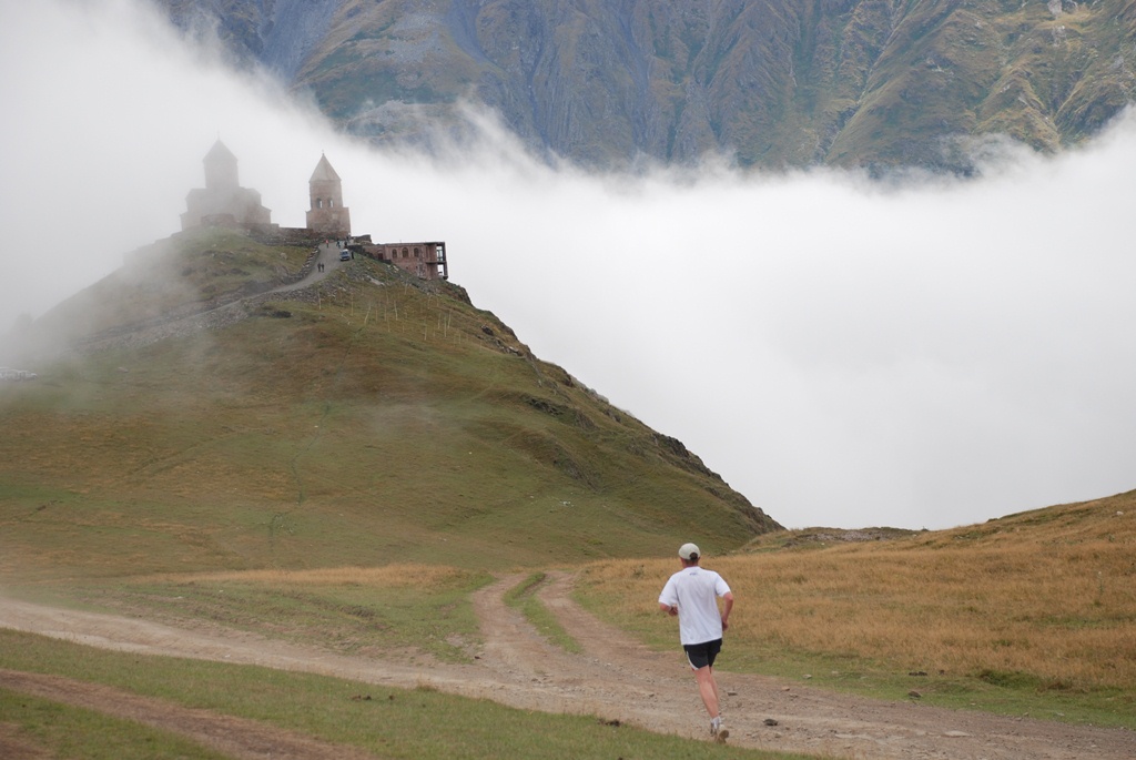 A marathon runner approaches Gergeti Trinity Church through drifting mountain fog.