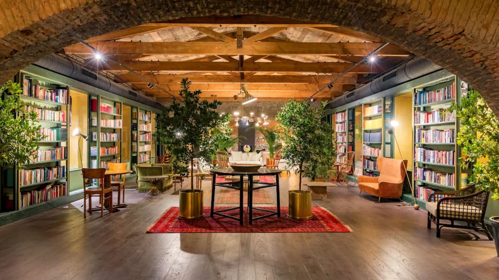 Library Bar — Просторный библиотечный зал с кирпичной аркой, пышной зеленью и рядами книжных полок.
