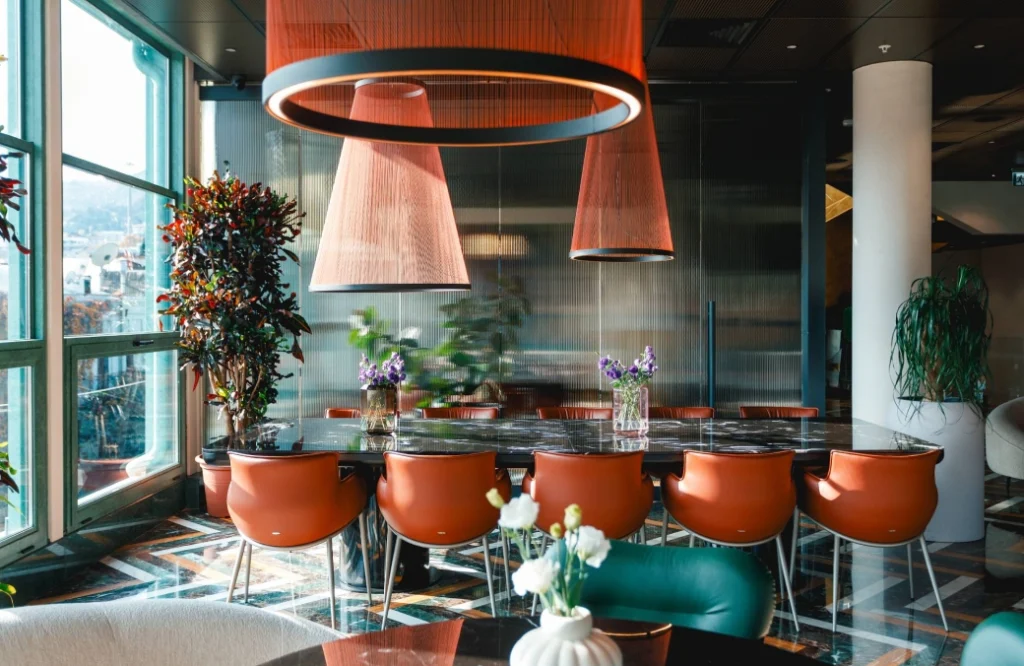 MOZAIKO — Dining area with orange upholstered chairs, round pendant lamps, and city view windows.