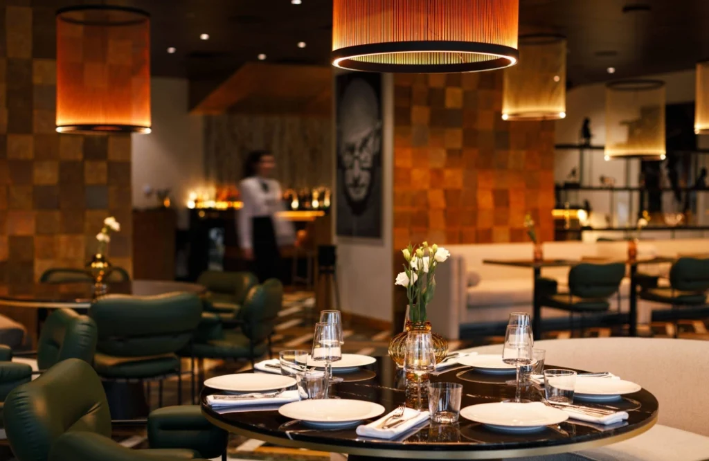 MOZAIKO — Elegant restaurant dining room with set tables, pendant lighting, and green upholstered chairs.