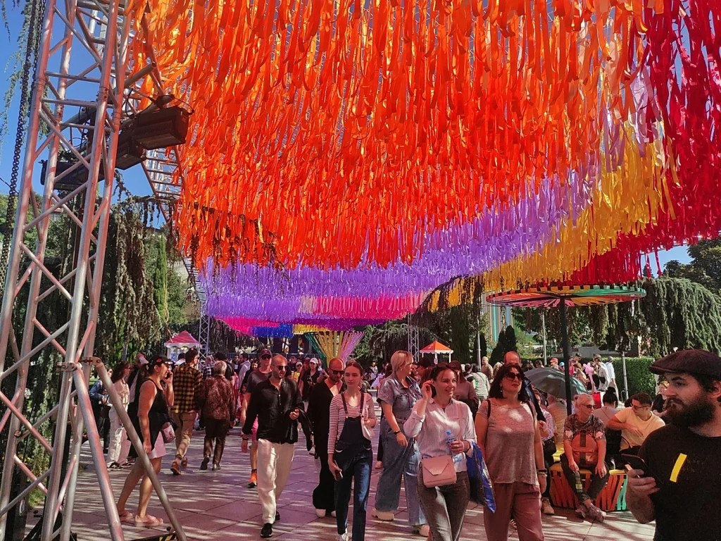 Festival-goers walk beneath colorful ribbon decorations at Tbilisoba in Rike Park.