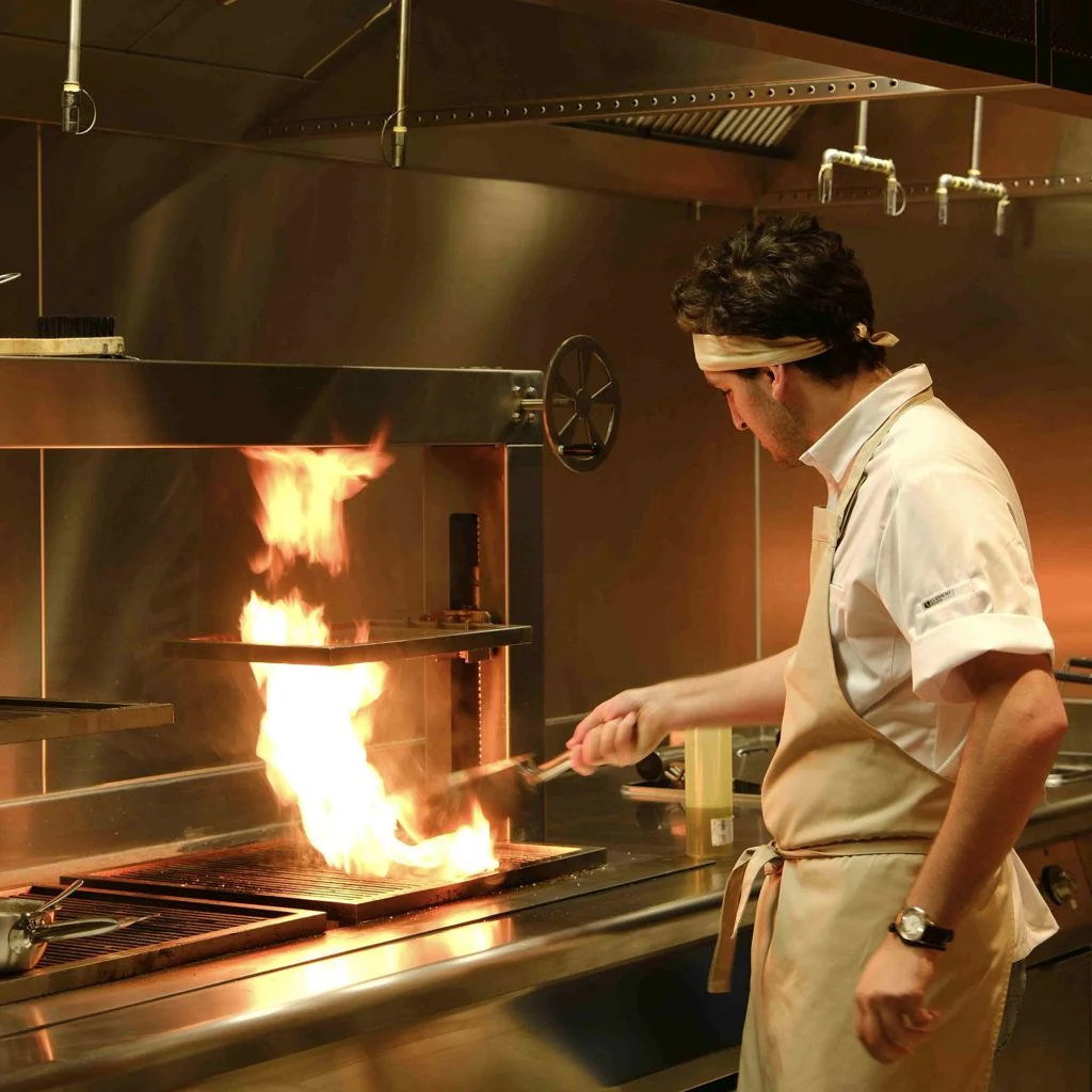 The Grill — Chef tending flames over an open grill in the restaurant's open kitchen.